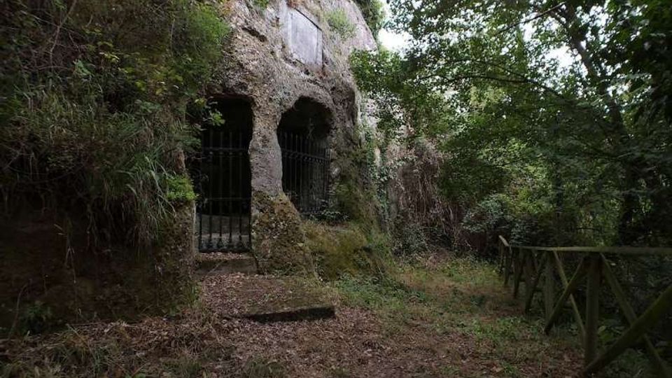 Ingresso scavato nella roccia lungo un sentiero boschivo, con due aperture ad arco chiuse da cancelli in ferro. Vegetazione fitta e alberi circondano l’area, con una staccionata in legno sul lato destro del percorso.