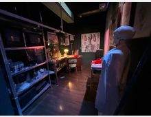 Interno di un piccolo laboratorio medico in penombra, con scaffalature metalliche piene di strumenti e contenitori, una scrivania illuminata da una lampada e poster anatomici alle pareti. Sul lato destro si vede un manichino e un lettino, in un ambiente con pavimento scuro e arredi essenziali.