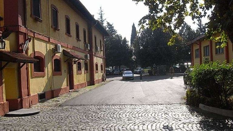 Piazzale esterno con pavimentazione in sampietrini, affiancato da un edificio a due piani color ocra con cornici rosse e finestre ad arco. Sullo sfondo si vedono alberi alti e un’area di parcheggio, con rami che proiettano ombre sull’acciottolato.