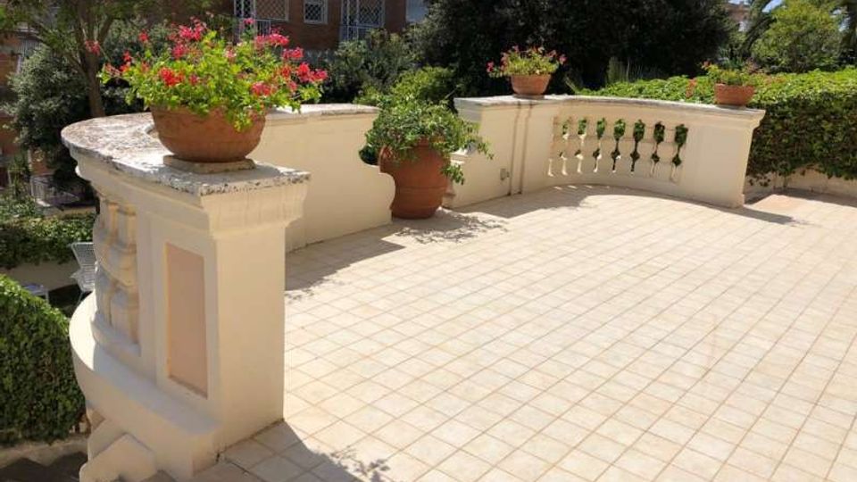 Terrazza esterna pavimentata con piastrelle chiare e parapetto curvo in muratura con balaustra; sui muretti sono appoggiati vasi in terracotta con fiori e piante. Intorno si vedono siepi e alberi, con un edificio residenziale sullo sfondo.
