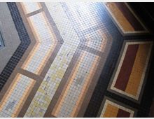 Pavimento interno in mosaico di piccole piastrelle quadrate, con motivi geometrici a bande e riquadri in tonalità grigio, beige, ocra e bordeaux. La composizione crea linee angolate e cornici che si incrociano lungo il corridoio.