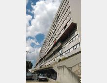 Grande edificio residenziale multipiano in cemento con lunga facciata lineare, file di finestre e ampi ballatoi incassati. In primo piano si vede una strada con auto parcheggiate, una rampa e scale in cemento, sotto un cielo parzialmente nuvoloso.