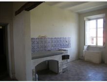 Interno di una cucina rustica e spoglia con pareti bianche, pavimento in piastrelle e una mensola in muratura rivestita di piastrelle decorative blu. A destra c’è una grande finestra con scuri aperti e, sotto, un lavello in muratura.