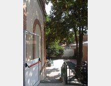 Cortile esterno pavimentato accanto a un edificio in muratura con arco in mattoni, con scala che scende e corrimano metallico. Alberi alti ombreggiano l’area; sullo sfondo si vedono un muretto, vegetazione e alcuni edifici bassi.