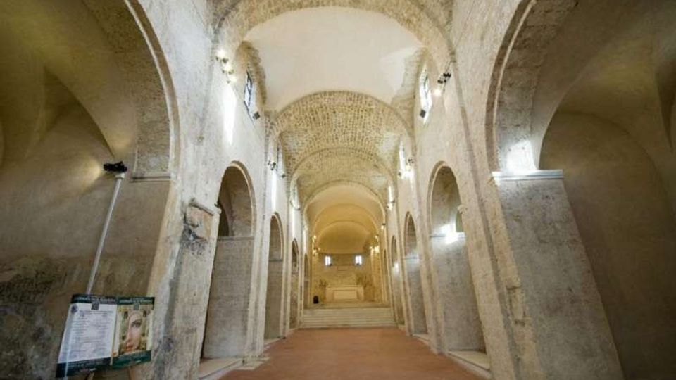 Interno di una chiesa o cripta in pietra con lunga navata centrale, alte arcate laterali e volte a botte illuminate da faretti. In fondo si vede una breve scalinata che conduce all’area dell’altare.