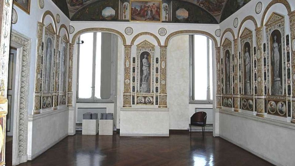 Sala storica con pavimento in legno scuro, pareti decorate da nicchie ad arco con figure dipinte e cornici dorate, e grandi finestre che illuminano l’ambiente. In alto, una fascia di affreschi corre sotto la volta, mentre l’arredo è minimo con una sedia e alcuni piccoli supporti.