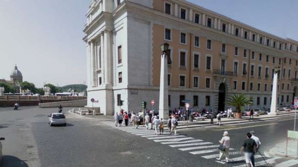 Incrocio urbano con ampio attraversamento pedonale davanti a un grande edificio storico in muratura chiara con facciata lunga e finestre regolari. Strada lastricata con traffico, marciapiedi e segnaletica; sullo sfondo si vede una cupola e un ponte in lontananza.