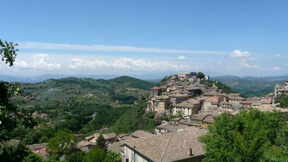 Veduta panoramica di un borgo collinare con case in pietra dai tetti in coppi, disposto lungo un crinale. Intorno si estendono colline verdi e campi coltivati sotto un cielo azzurro con nuvole all’orizzonte.