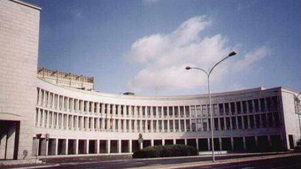 Edificio moderno a più piani con facciata curva e lunga serie di finestre verticali ripetute, affacciato su una strada ampia. In primo piano si vedono un’aiuola centrale e alti lampioni, con cielo azzurro sullo sfondo.