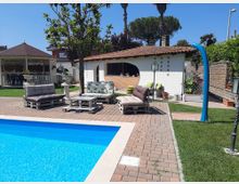 Cortile di una villa con piscina rettangolare azzurra in primo piano e pavimentazione in mattoni. Sullo sfondo ci sono divani da esterno su pallet, un gazebo bianco e un piccolo edificio con tetto in coppi e veranda, circondati da prato, piante e muri di confine.