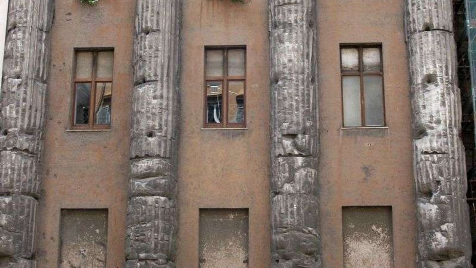 Facciata di un edificio storico con alte colonne scanalate in pietra scura, rovinate e ricomposte, che incorniciano una parete intonacata con finestre rettangolari. Alla base si vedono grandi blocchi irregolari e una balaustra lungo il marciapiede su una strada urbana.