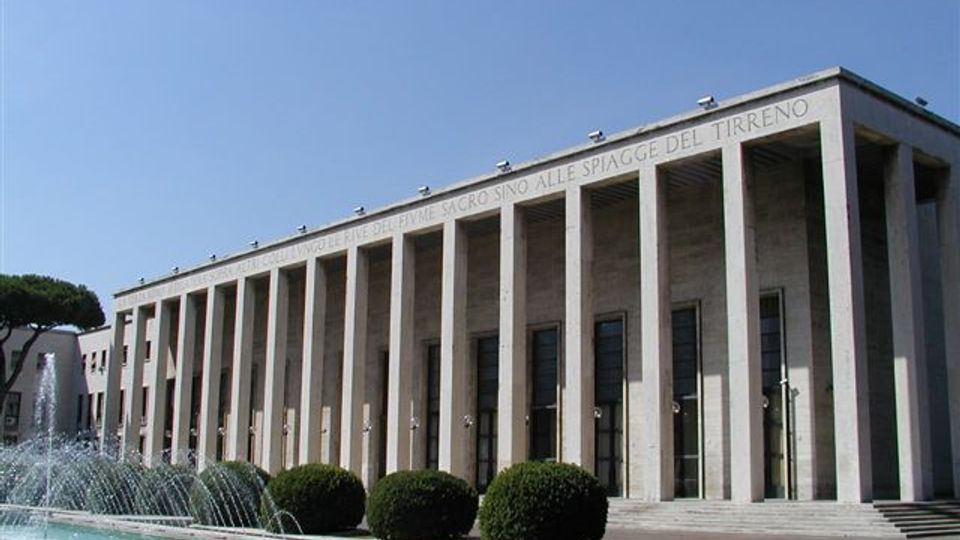 Edificio monumentale in stile razionalista con lunga facciata in pietra scandita da alte colonne rettangolari e grandi finestre. Davanti si trovano una fontana con getti d’acqua, una piazza aperta e siepi tondeggiantI sotto un cielo sereno.