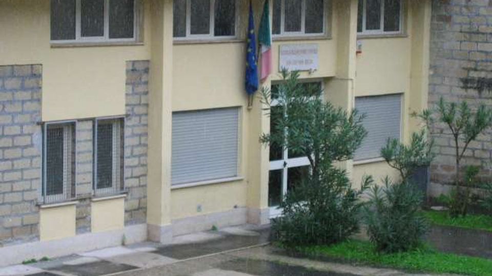 Edificio pubblico a due piani con facciata giallo chiaro e parti in muratura grezza, tetto in tegole rosse e finestre con serrande metalliche. All’ingresso con porta vetrata sono presenti bandiere e un cartello, davanti a un piccolo giardino con arbusti e un piazzale asfaltato bagnato.