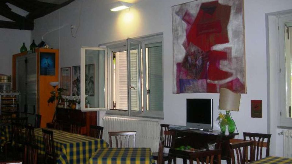 Sala interna tipo trattoria con tavoli e sedie in legno coperti da tovaglie a quadri giallo‑blu, pareti chiare e soffitto con travi scure a vista. Sulla parete ci sono una finestra aperta con persiane, un termosifone, un grande quadro astratto e un piccolo angolo con televisore e lampada.