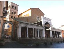 Facciata di una chiesa in mattoni con portico colonnato e grande arco d’ingresso, con rosone circolare sulla parete frontale. Sul lato si vede un campanile a più livelli con aperture ad arco e, davanti, una recinzione in ferro lungo la piazza.