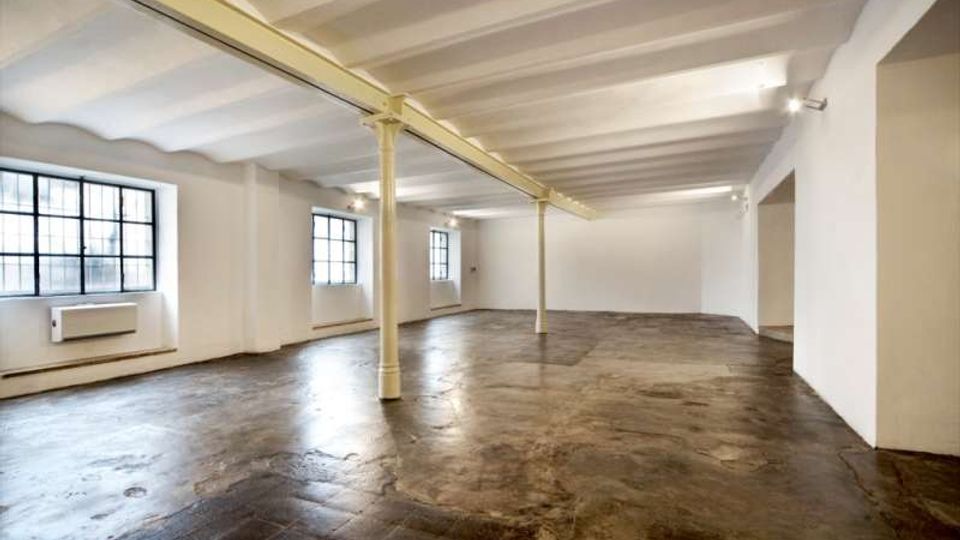 Ampio spazio interno vuoto in stile loft industriale, con pavimento in cemento, soffitto con travi a vista e colonne metalliche centrali. Lungo una parete ci sono grandi finestre a griglia che illuminano l’ambiente, con pareti bianche e luci a parete.
