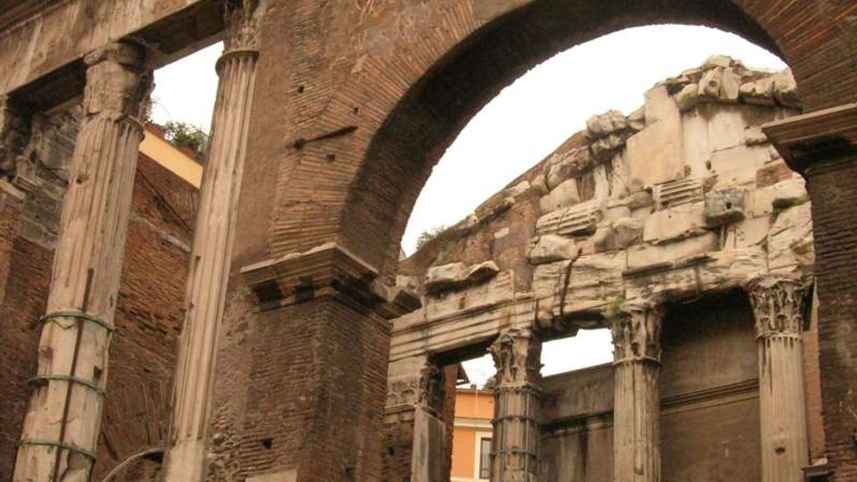 Rovine di una struttura romana con grande arco in mattoni e colonne in pietra scanalate, alcune con capitelli decorati. Sullo sfondo si vedono muri parzialmente crollati e un edificio moderno, con recinzioni metalliche in primo piano.
