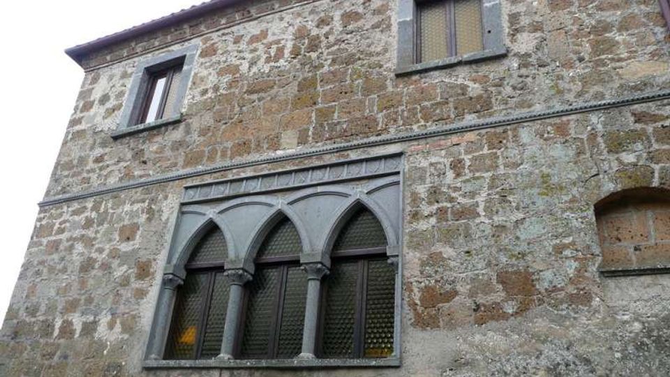 Facciata di un edificio storico in pietra con intonaco consumato, due finestre rettangolari al piano superiore e una grande finestra a tre archi ogivali con colonnine al piano inferiore. Cornici e decorazioni in pietra grigia evidenziano l’architettura.