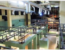 Interno di un grande edificio industriale riconvertito, con passerelle e ringhiere metalliche sopraelevate e una fitta rete di condotti e tubazioni d’aerazione a soffitto. Lo spazio è diviso da pareti verde chiaro e pilastri, con aree espositive illuminate al livello inferiore.
