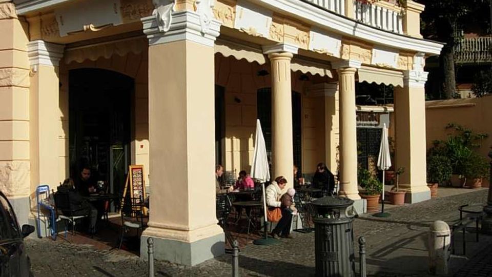 Portico di un edificio storico color crema con grandi colonne e decorazioni in stucco, affacciato su una piazzetta con pavimentazione in sampietrini. Sotto il portico si vede l’area esterna di un bar con tavolini, sedie e ombrelloni chiusi, circondata da piante in vaso e paletti lungo il marciapiede.