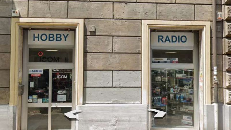 Facciata di un edificio in pietra con due ingressi vetrati affiancati, con insegne “HOBBY” e “RADIO” sopra le porte e vetrine che mostrano prodotti elettronici. Davanti sul marciapiede sono parcheggiati scooter e moto lungo la strada.