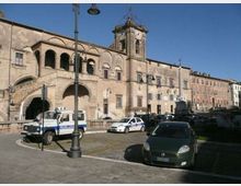 Piazza urbana con pavimentazione in pietra e auto parcheggiate, affacciata su un grande edificio storico in muratura con porticato ad archi, scala esterna e torre campanaria con orologio. Lampioni in ferro e facciate di palazzi allineati lungo la strada sotto un cielo sereno.