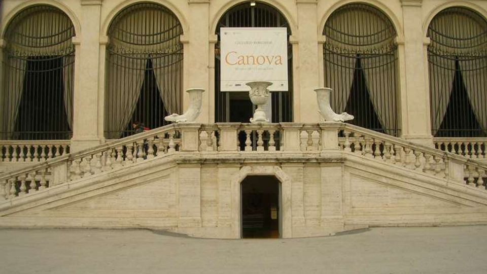 Facciata di un edificio storico con grandi finestre ad arco e grate in ferro, affacciata su una scalinata simmetrica in pietra con balaustra. Al centro si vede un ingresso sottostante e un grande cartellone espositivo con la scritta “Canova”.