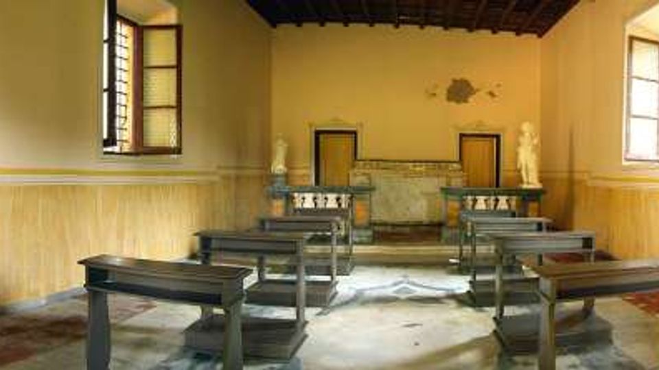Interno di una piccola aula con pareti gialle e zoccolatura in finto marmo, file di banchi in legno e pavimento in parte rovinato. In fondo si vede una pedana con cattedra e una parete decorata, con finestre laterali e soffitto scuro a travi.