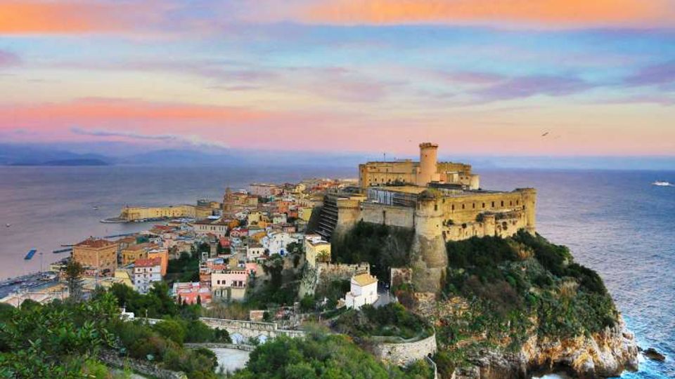 Veduta panoramica di un borgo costiero con case addossate e un grande castello fortificato con torre cilindrica su un promontorio roccioso. Il mare circonda la scogliera e sullo sfondo si vede l’orizzonte sotto un cielo al tramonto.