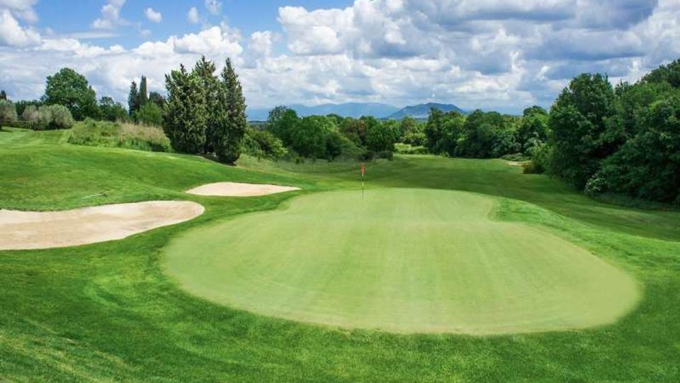 Campo da golf con ampio green in primo piano e bandierina al centro, circondato da fairway ondulati e bunker di sabbia sulla sinistra. Sullo sfondo si vedono alberi, colline e un cielo con nuvole sparse.