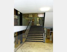 Atrio interno di un edificio con ampia scala centrale in pietra scura e corrimano in metallo, pavimento in marmo chiaro e soffitto con pannelli e illuminazione al neon. In fondo alla scalinata si vede una statua su piedistallo e pareti decorate con disegni e quadri.