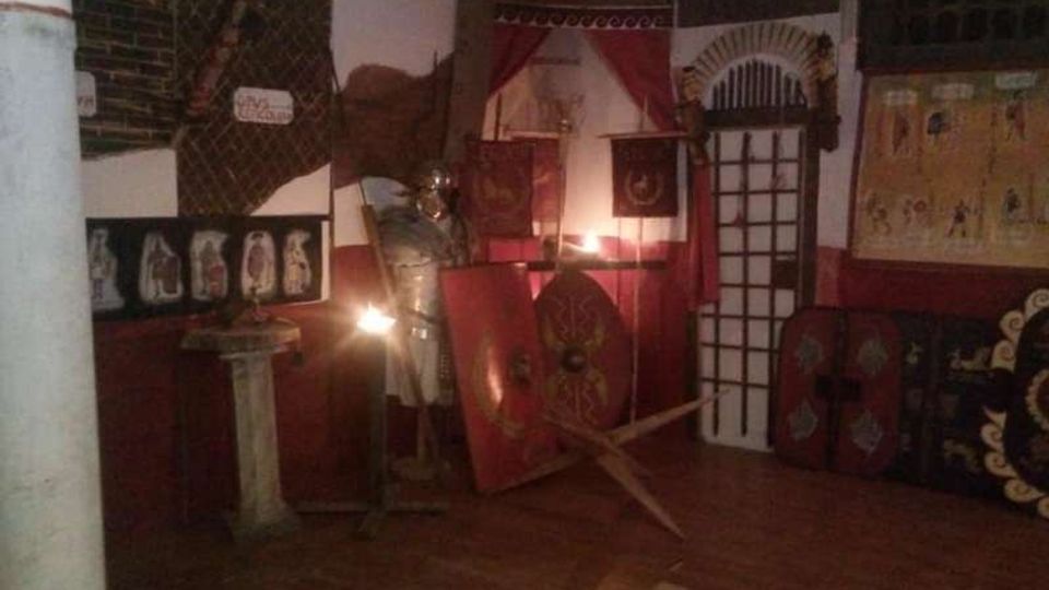 Interno di una sala espositiva a tema romano, con pareti in mattoni e pannelli rossi decorati, scudi e armature in mostra e un piccolo altare o piedistallo su pavimento in legno. Sul fondo si vedono drappi rossi, una grata a finestra e teche o pannelli con oggetti e illustrazioni.