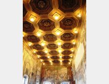Soffitto a cassettoni ottagonali in legno, illuminato da numerose luci calde, in una lunga sala decorata. Le pareti laterali e di fondo sono rivestite da grandi affreschi con scene figurative e archi dipinti.