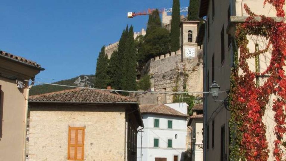 Strada stretta in un borgo con case in pietra e facciate chiare, tetti in coppi e auto parcheggiate lungo il margine. Sullo sfondo si vede una fortificazione su una collina con cipressi e una torre con orologio, sotto un cielo sereno.