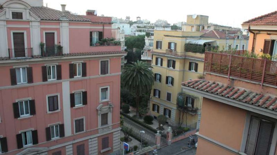 Vista dall’alto di un quartiere urbano con palazzi residenziali color rosa e giallo, tetti in coppi e persiane scure. Tra gli edifici si vede una strada e un piccolo giardino con una grande palma e vegetazione.