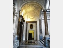 Interno di un edificio storico con atrio a volta decorata a cassettoni e stucchi, colonne in pietra e cornici in marmo attorno a una porta centrale. Pavimento in marmo con motivo geometrico e scalinate laterali sotto archi ornamentali.