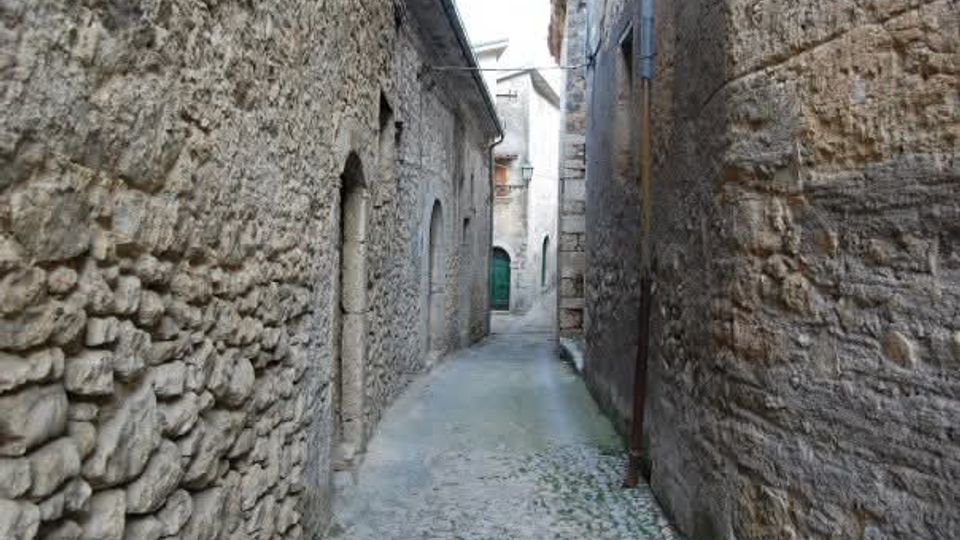 Vicolo stretto in un borgo storico, con pavimentazione in ciottoli e alte pareti in pietra grezza ai lati. In fondo si intravedono archi e porte di edifici in muratura lungo la strada curva.