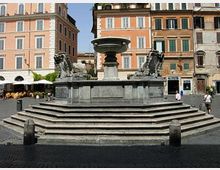 Grande fontana in pietra con vasca centrale su base a gradoni e sculture laterali, collocata in una piazza lastricata. Sullo sfondo si vedono alti edifici storici con facciate color ocra e persiane, oltre a un’area con tavolini all’aperto.