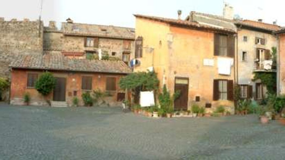 Piazza lastricata in pietra in un borgo storico, circondata da case in muratura dai toni ocra e rosa con persiane, balconi e vasi di piante. Sullo sfondo si vede un arco in pietra inserito nelle mura, che conduce a una strada oltre il centro abitato.