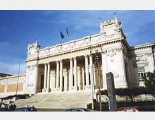 Facciata monumentale di un edificio neoclassico con ampio portico a colonne e lunga scalinata in pietra che conduce all’ingresso. La struttura chiara presenta decorazioni e frontone con bandiere, affacciata su una strada con auto in primo piano e cielo sereno sullo sfondo.