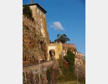 Fortezza in pietra costruita su una scogliera rocciosa, con edificio alto e mura merlate che seguono il profilo del promontorio. In primo piano si vede un arco d’ingresso giallo collegato a una strada in salita, con vegetazione sulle rocce e cielo azzurro sullo sfondo.