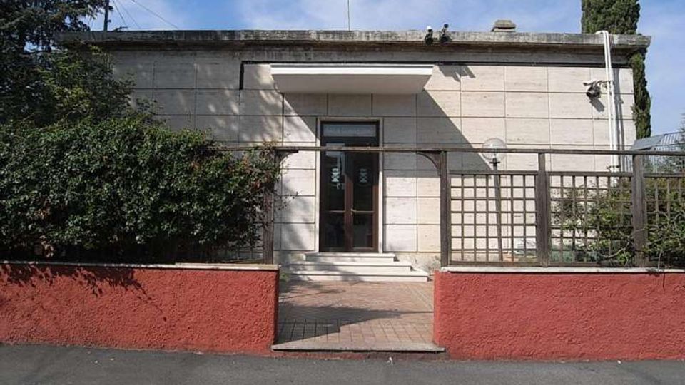 Facciata di un piccolo edificio moderno a un piano, rivestito in pietra chiara, con ingresso centrale a porta vetrata sotto una pensilina. Davanti c’è un camminamento con muretto basso rosso e una grata in legno, circondato da siepi e alberi.