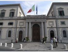 Facciata di un edificio istituzionale in stile neoclassico, con ingresso centrale ad arco affiancato da colonne e grandi portoni in legno. In cima sventolano le bandiere italiana e dell’Unione Europea, davanti a un piazzale lastricato con dissuasori in pietra.