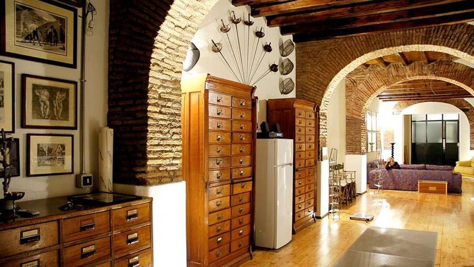Interno di un appartamento in stile loft con grandi archi in mattoni a vista, soffitto con travi in legno e pavimento in parquet. Lungo il corridoio ci sono alte cassettiere in legno e arredi moderni, con un’area soggiorno sullo sfondo.