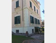 Facciata angolare di una villa a più piani intonacata in rosa con cornici bianche, persiane verdi e finestre alte con dettagli decorativi. In primo piano si vedono un vialetto in pietra con motivo circolare, un prato e un vaso su piedistallo vicino a un’area con pergolato.