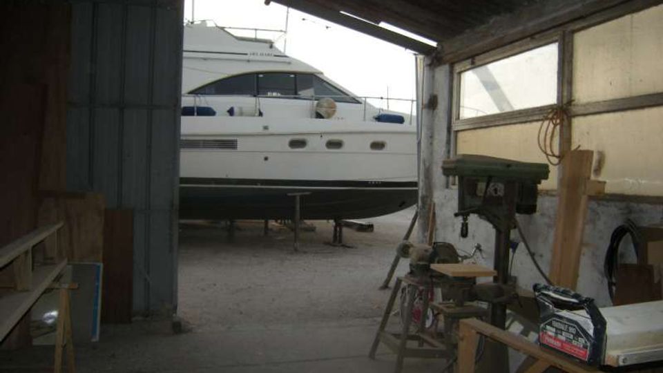 Interno di un capannone rimessaggio con tetto in lamiera e pareti in legno e metallo, aperto verso un piazzale. All’esterno, un grande yacht bianco su cavalletti è visibile attraverso l’apertura, mentre all’interno si notano banchi da lavoro e attrezzi da falegnameria.