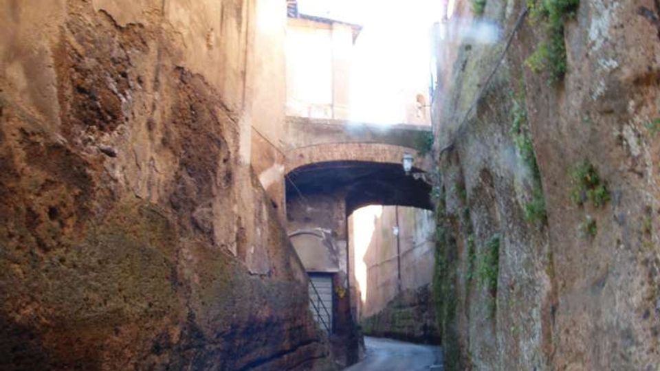 Vicolo stretto e in pendenza tra alte pareti di roccia e muratura, con strada asfaltata che curva in fondo. Un piccolo arco in pietra collega gli edifici sopra la via e sulle pareti si notano segni di erosione e vegetazione.