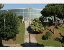 Edificio moderno a pianta semicircolare con ampia facciata vetrata, situato in cima a una scalinata centrale che risale un pendio erboso. Ai lati della scalinata ci sono alberi e cespugli, con lampioni e recinzioni lungo l’ingresso.
