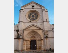Facciata in pietra di una chiesa storica con grande rosone circolare centrale e portale ad arco ogivale con colonne laterali e porta in legno scuro. Sopra il rosone si vede un piccolo oculo e un frontone triangolare, con elementi decorativi lungo la cornice sotto un cielo azzurro.