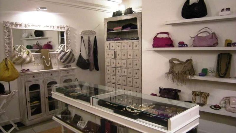 Interno di una boutique di accessori con pareti bianche, scaffali a vista con borse e piccoli oggetti esposti e mobili in legno chiaro. Al centro ci sono vetrine in vetro per gioielli, con cassettiere e specchi decorativi lungo le pareti.